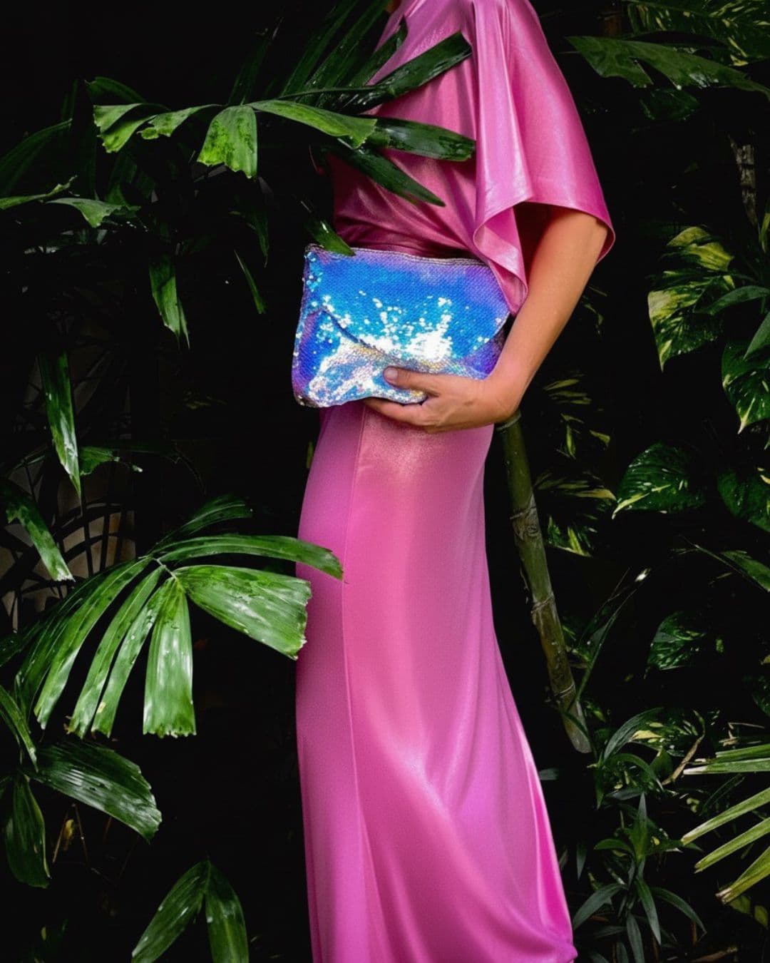 Person in a pink dress holding a silver sequin clutch against a dark green leafy background