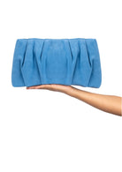 A blue pleated suede clutch bag held by a person's hand against a plain white background.