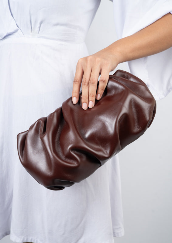 Bordeaux Brown textured clutch held by a person wearing a white outfit on a light gray background